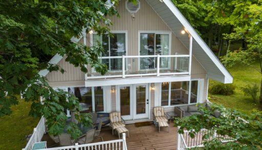 A beige cottage with white trim. On the lower level deck, two brown lounge chairs.