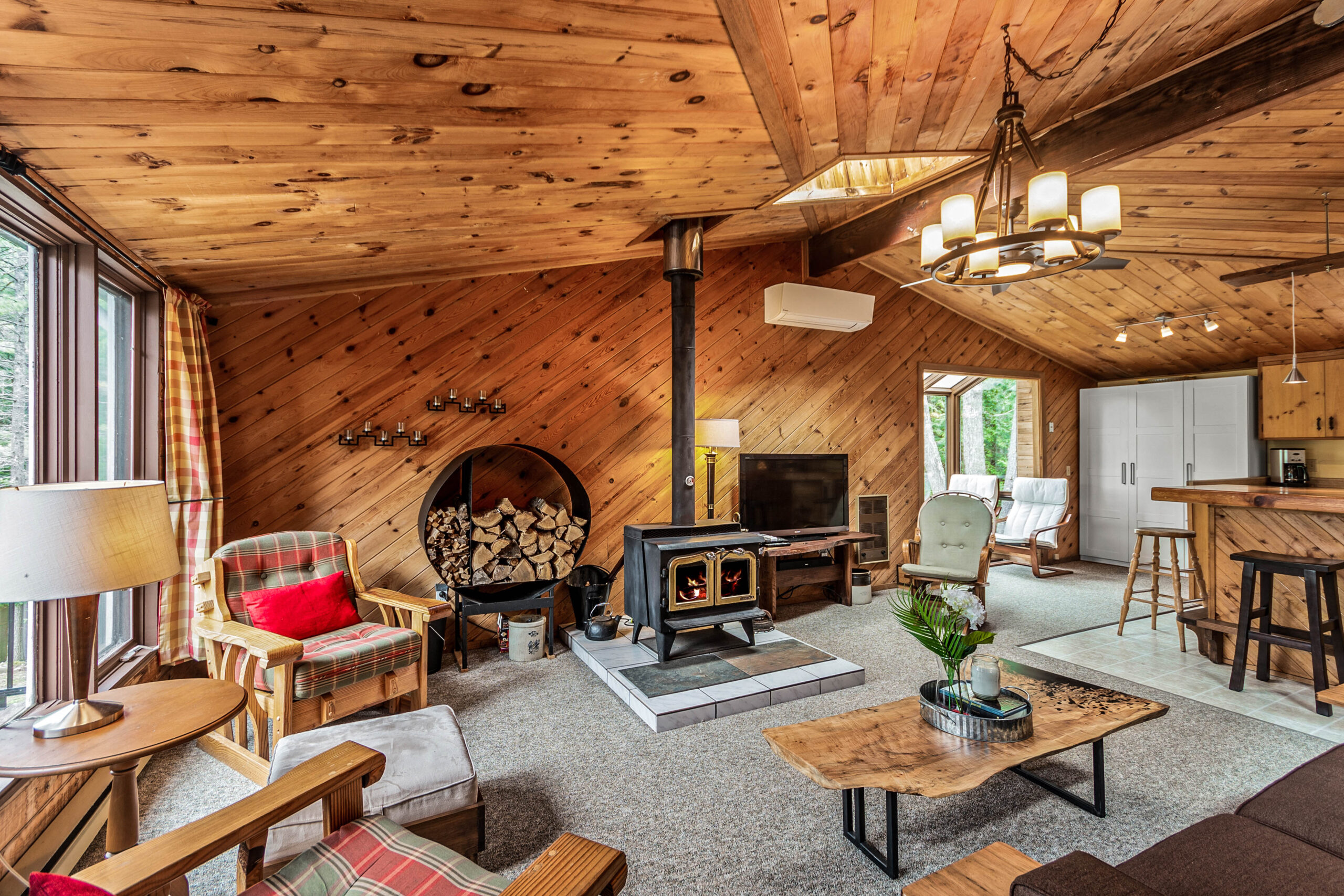 A wood stove in a wood-paneled room. Logs are stored in a round shelf on the left while a TV is on the right