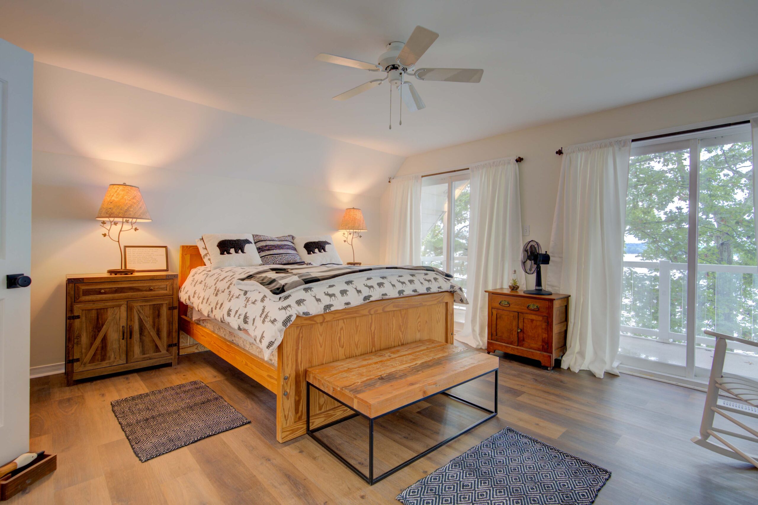 A bed on a large wood bed frame with a small wood bench at the end. To the left, bright windows that face the lake