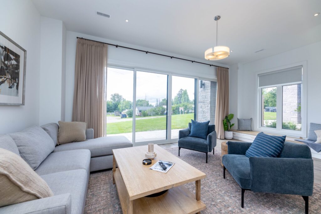 A white couch faces a light wood coffee table and two blue chairs. To the left, bright windows face a grassy lawn