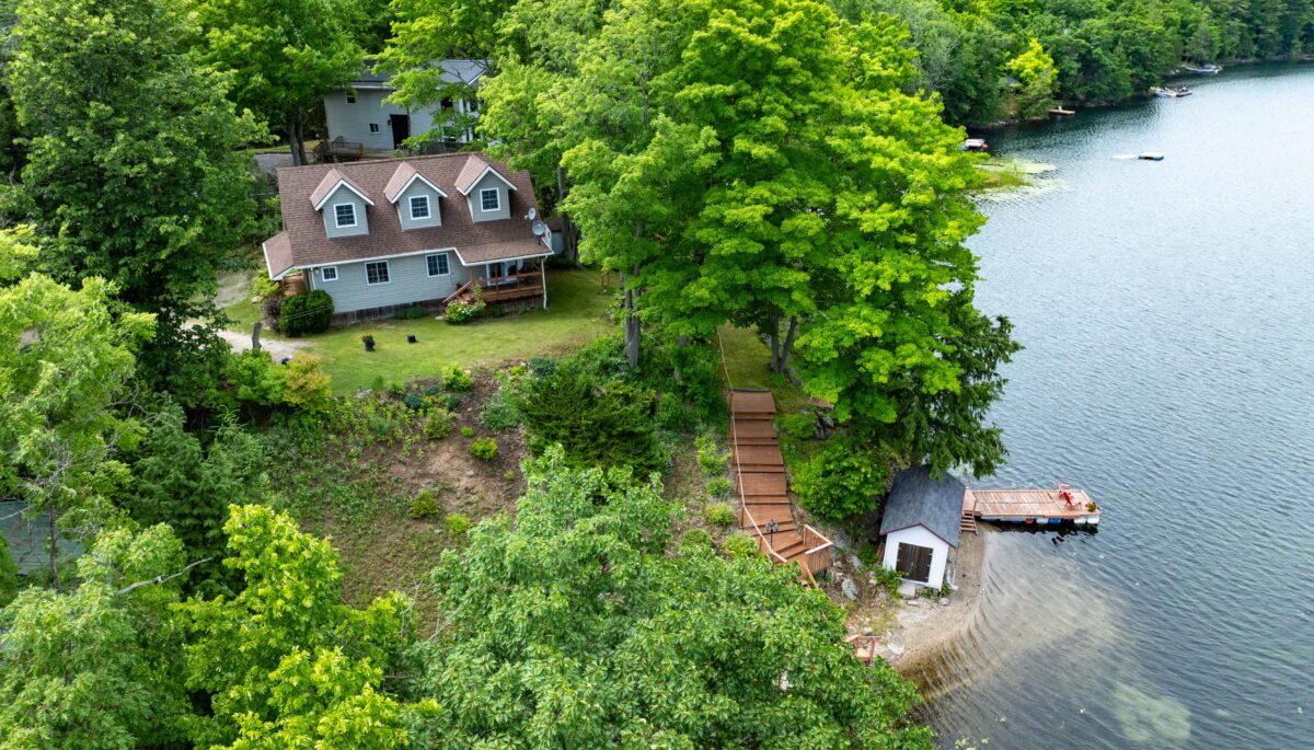 A paneled-cottage with three upper windows is set back from the lake on a lush green property