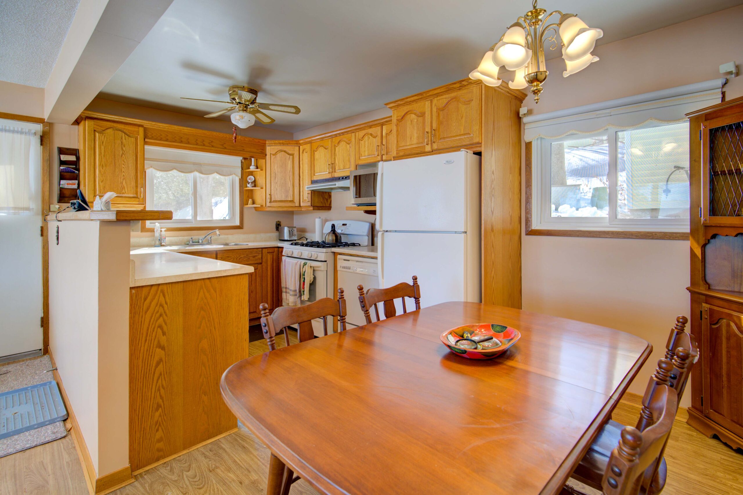 A wood dining table with wood chairs is adjacent to a wood kitchen with a white fridge
