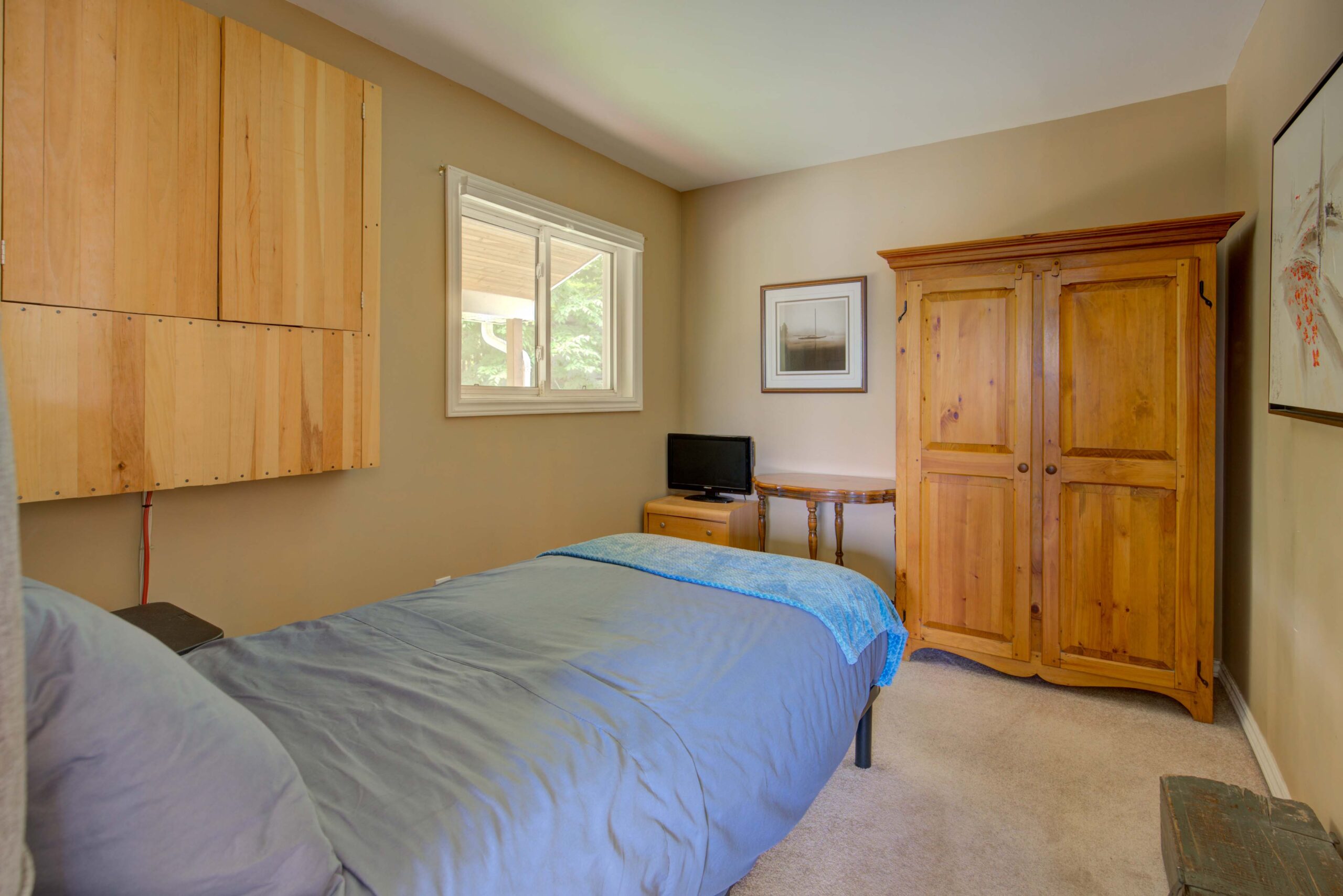 A blue bed in a beige bedroom faces a wood armoire