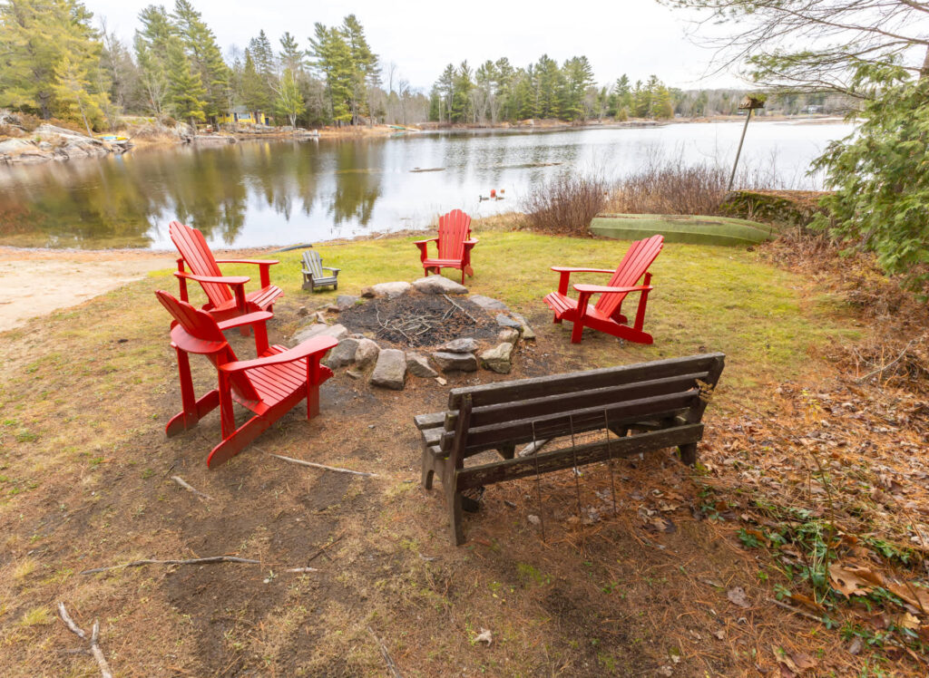 Four red Muskoka chairs and a wood bench around a lakeside fire pit
