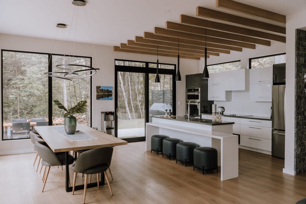 A modern kitchen with a white island and a pale wood dining table. Wood panelling is dotted along the ceiling