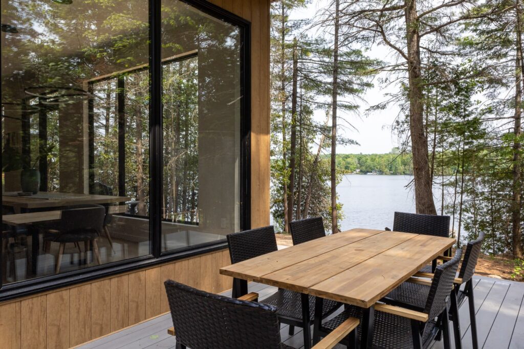 A wood outdoor dining table with black chairs outside a glass-walled cottage