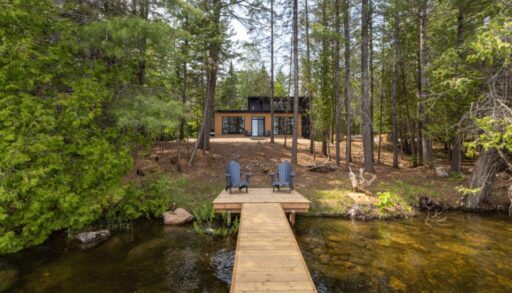 A black cottage is set back from the lake. In front, a pale wood dock with two blue Muskoka chairs