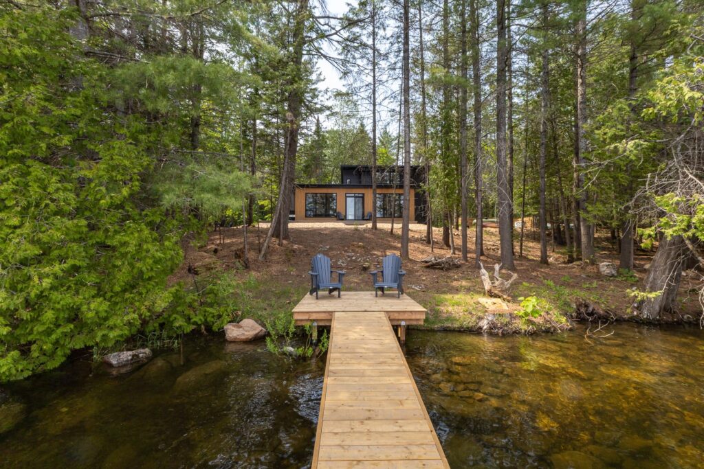 A black cottage is set back from the lake. In front, a pale wood dock with two blue Muskoka chairs