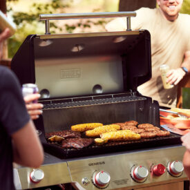 A variety of meats and corn grilling on a barbecue with people standing around it
