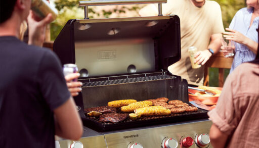 A variety of meats and corn grilling on a barbecue with people standing around it