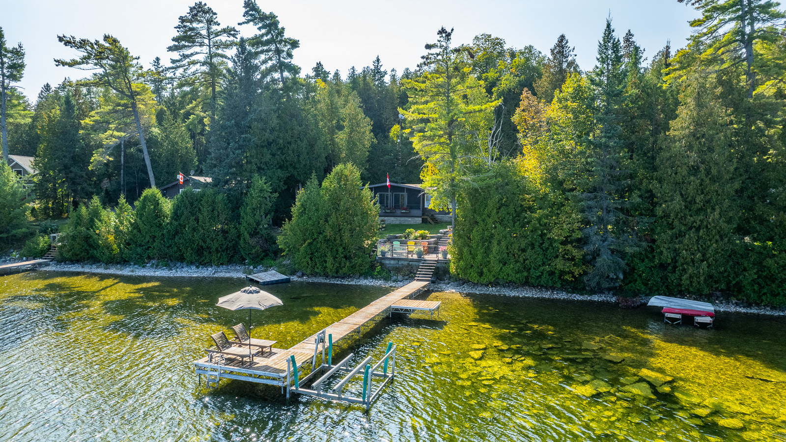 A brown paneled cottage set back in the trees. By the shoreline, a long dock juts out into the water