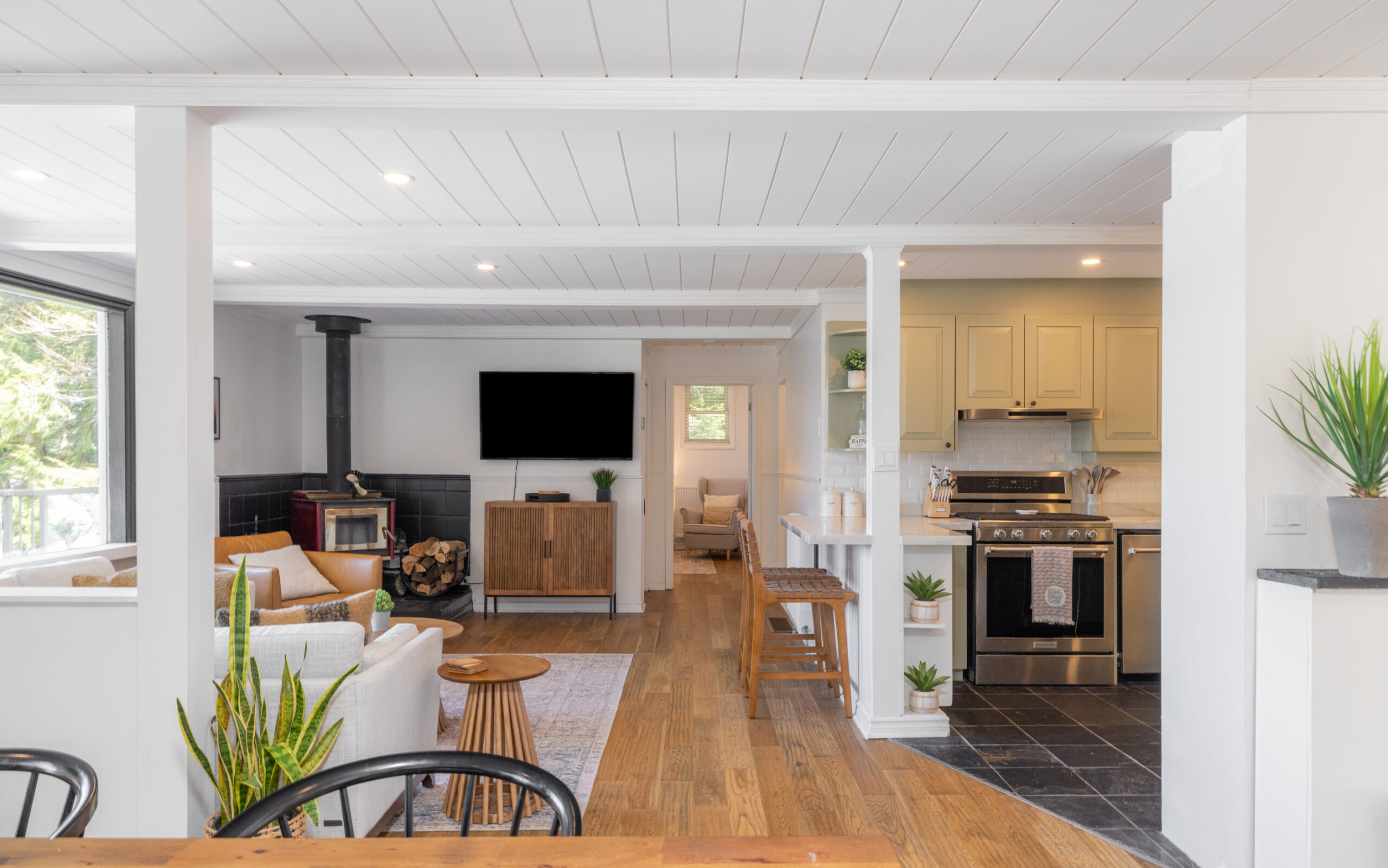 A spacious living area with a white couch facing a wood stove and TV. To the right, a kitchen