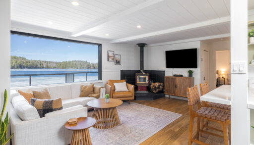 A white couch next to a bright lake-facing window faces a wood stove