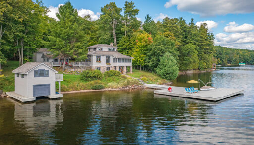 Scenic waterfront view of a Lake of Bays cottage with a two-storey boathouse, dock, and lush surrounding forest under a blue sky