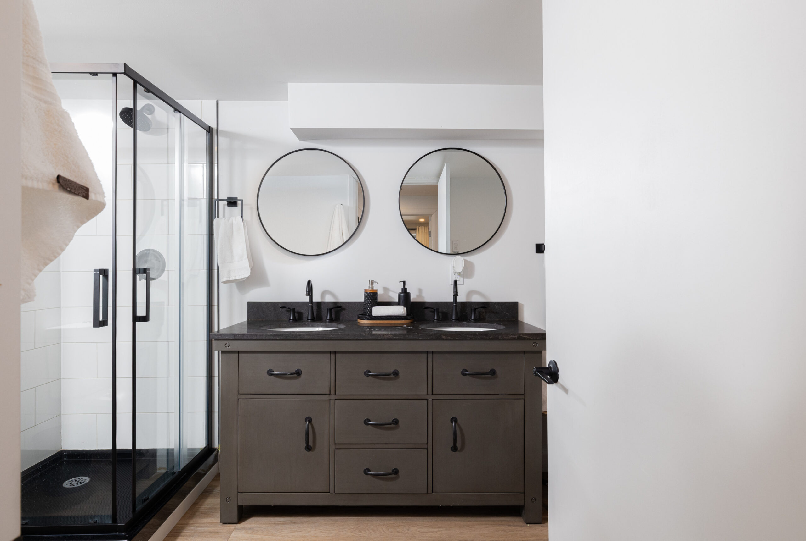 A dark grey vanity with two round mirror hanging above. To the left, a glass walk-in shower