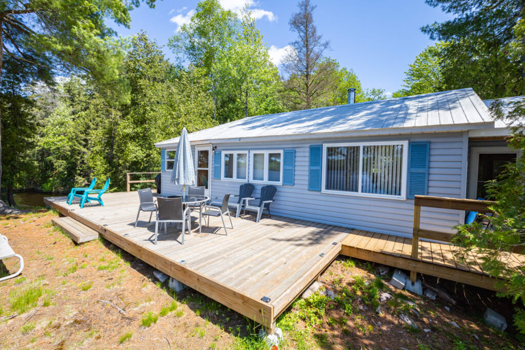 Exterior of light blue cottage with deck, patio set, and surrounding forest