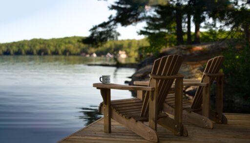Two Adirondack chairs sit on a cottage dock, facing a calm Muskoka lake with cabins in view on a sunny summer morning. A coffee tin on one arm reads "Life is better at the Cottage