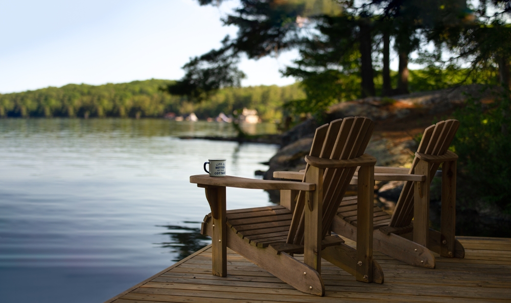 Two Adirondack chairs sit on a cottage dock, facing a calm Muskoka lake with cabins in view on a sunny summer morning. A coffee tin on one arm reads "Life is better at the Cottage