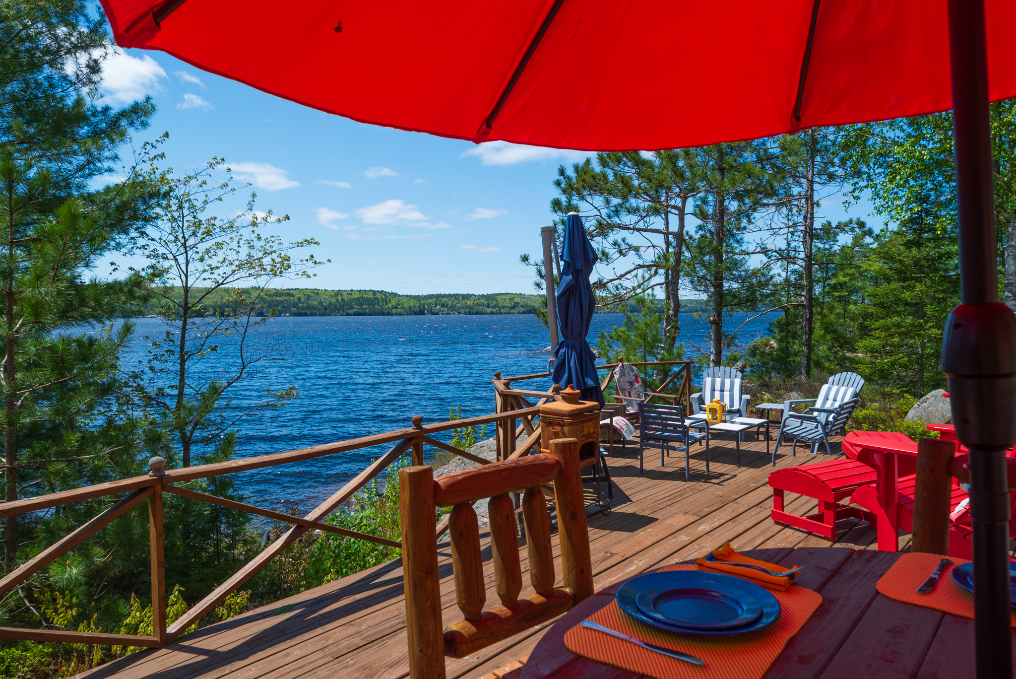 Muskoka chairs and table set under a red umbrella on a sunny dock overlooking Mazinaw Lake