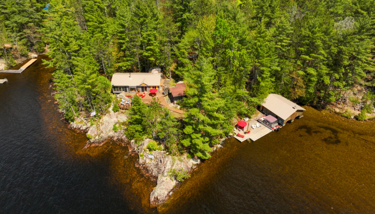 Drone shot capturing the shoreline, dense forest, sandy beach, and boathouse of 890 Mazinaw Lake