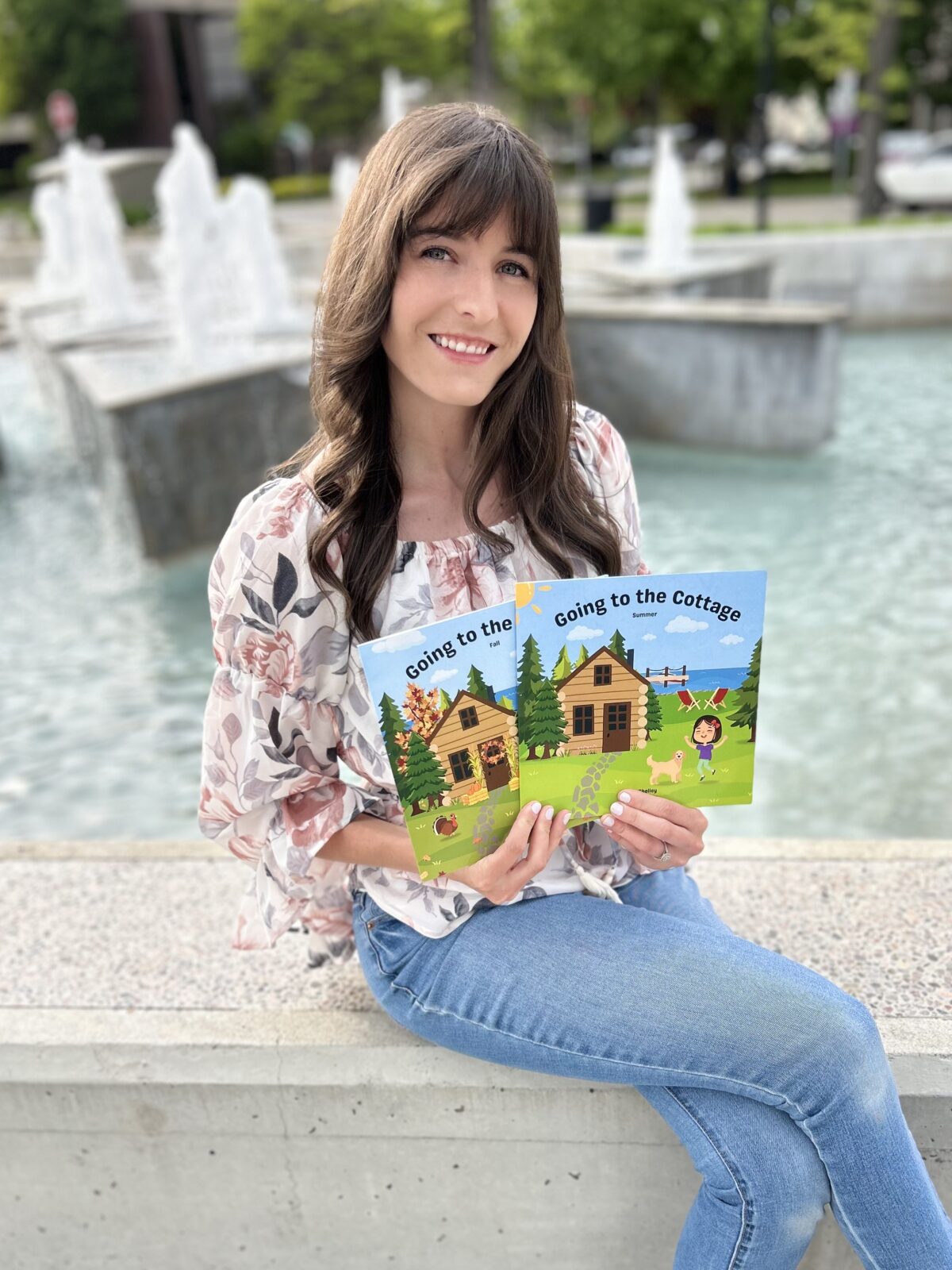 A young woman sitting in front of a fountain posing with children's books that she wrote called Going to the Cottage.