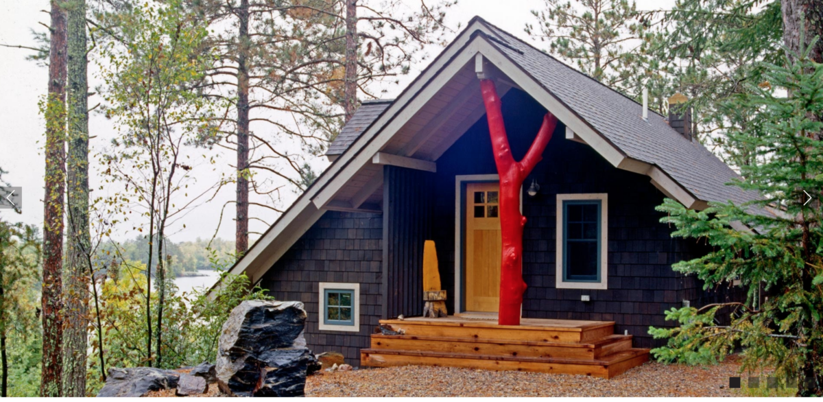 The cabin of cabinologist Dale Mulfinger, pictures with dark siding and a red tree trunk accenting the front porch.