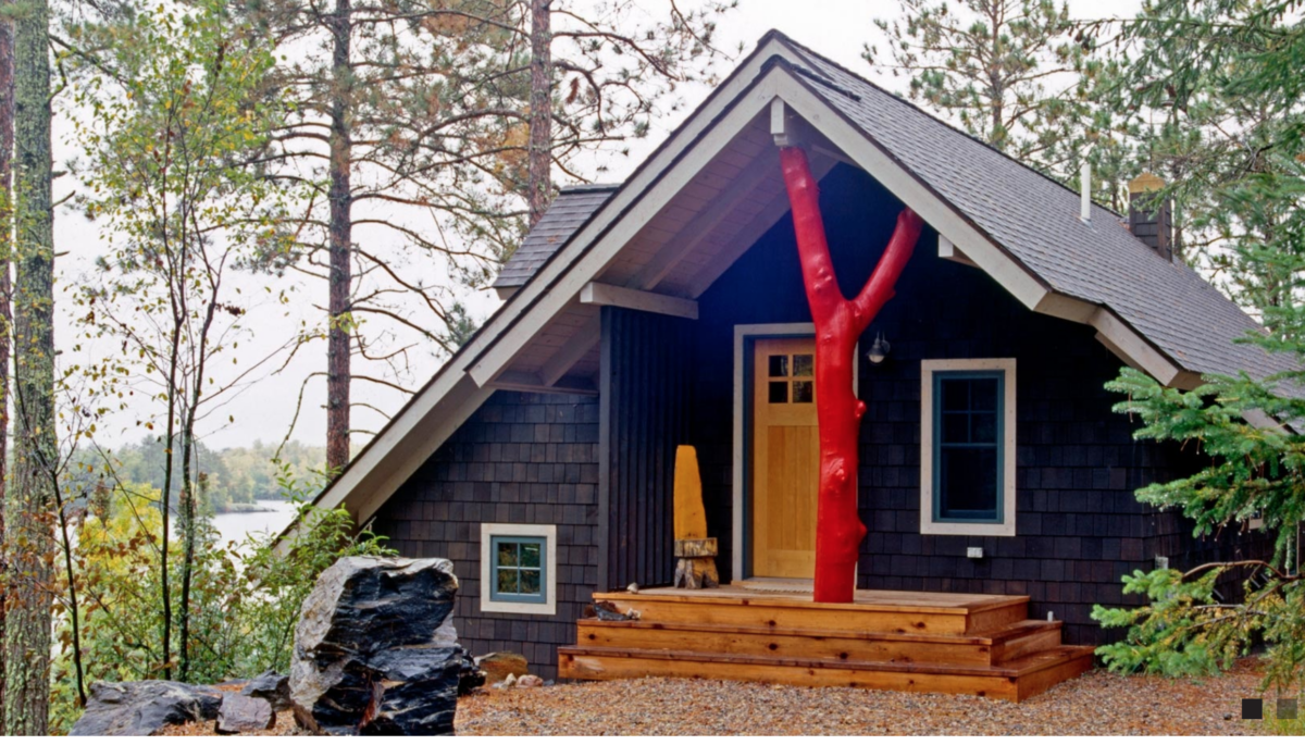 The cabin of cabinologist Dale Mulfinger, pictures with dark siding and a red tree trunk accenting the front porch.