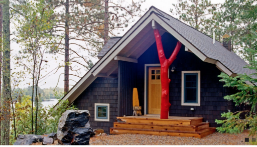 The cabin of cabinologist Dale Mulfinger, pictures with dark siding and a red tree trunk accenting the front porch.