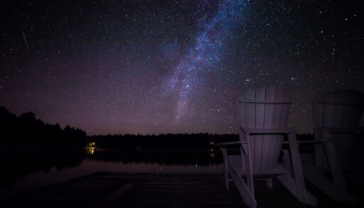 Muskoka chairs on a dock at night with the Milky Way in the background. Light pollution