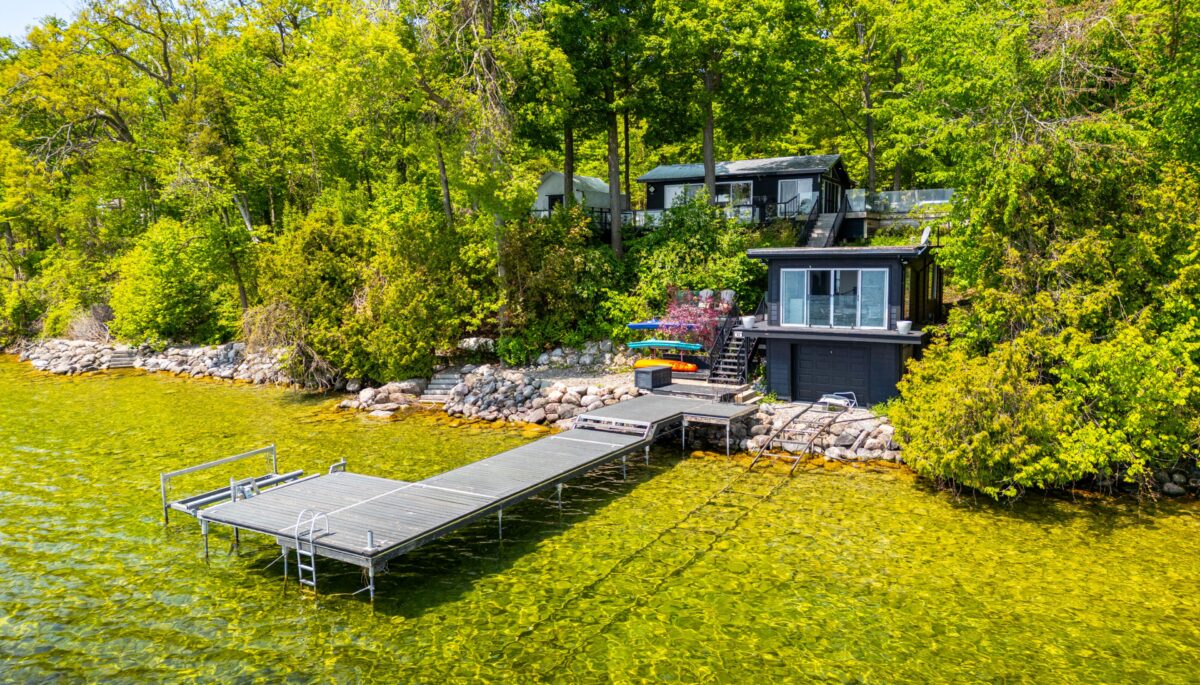 A black paneled cottage with a long deck that juts out into green-blue water
