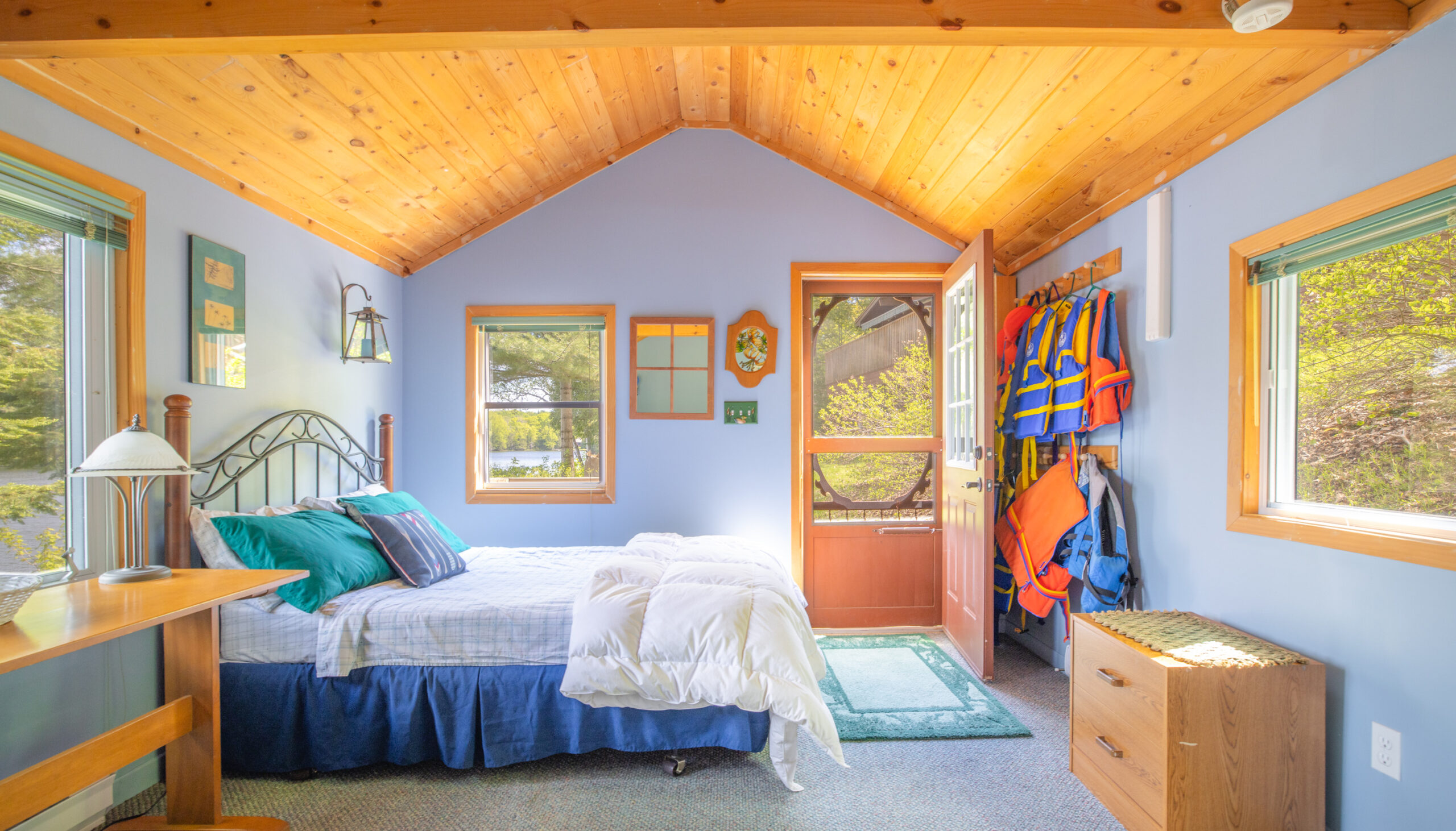 A blue bedroom with wood slanted ceilings. A bed faces a wall of lifejackets