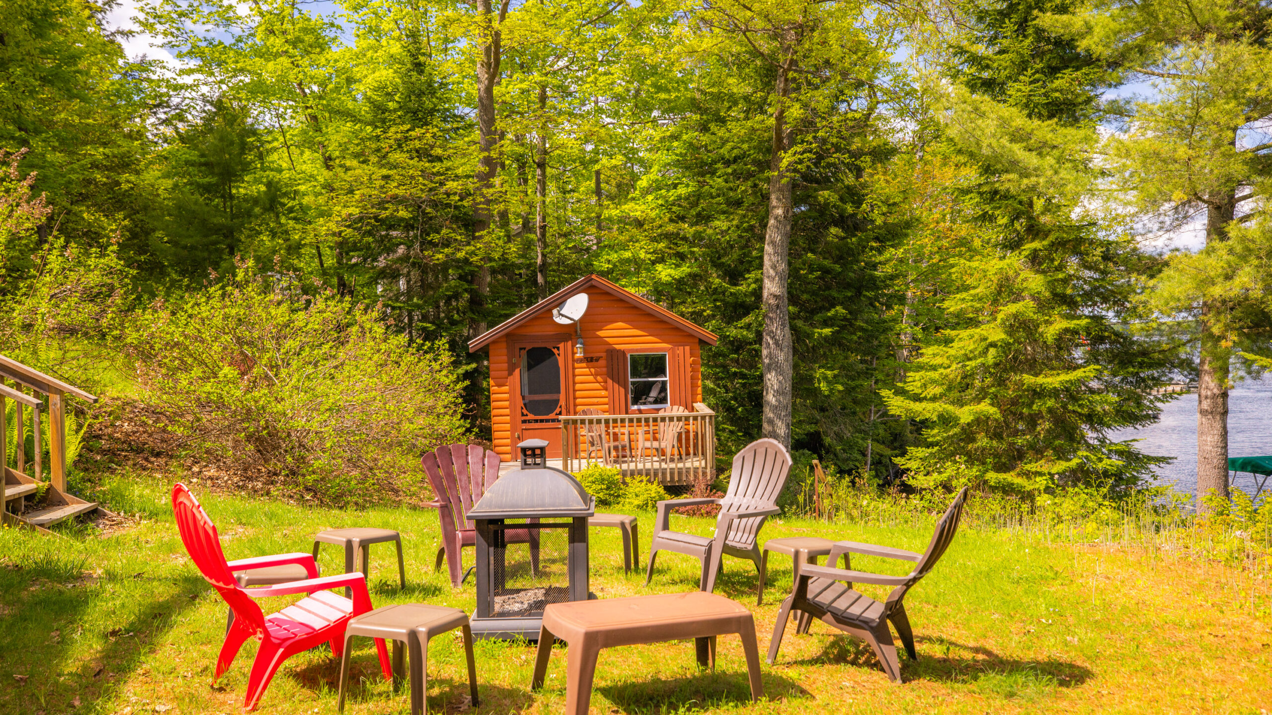Muskoka chairs around a fire pit. In the distance, a small wood dwelling nestled in the trees