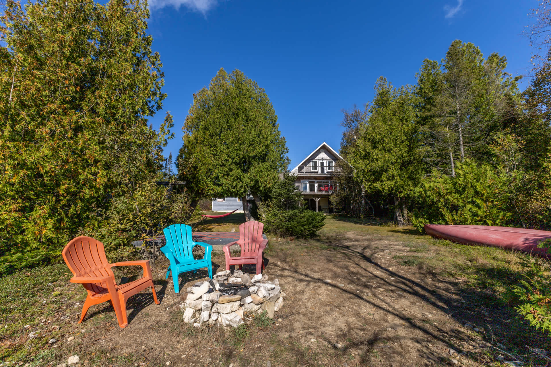 A stone fire pit with colourful Muskoka chairs around it. In the distance, a large cottage
