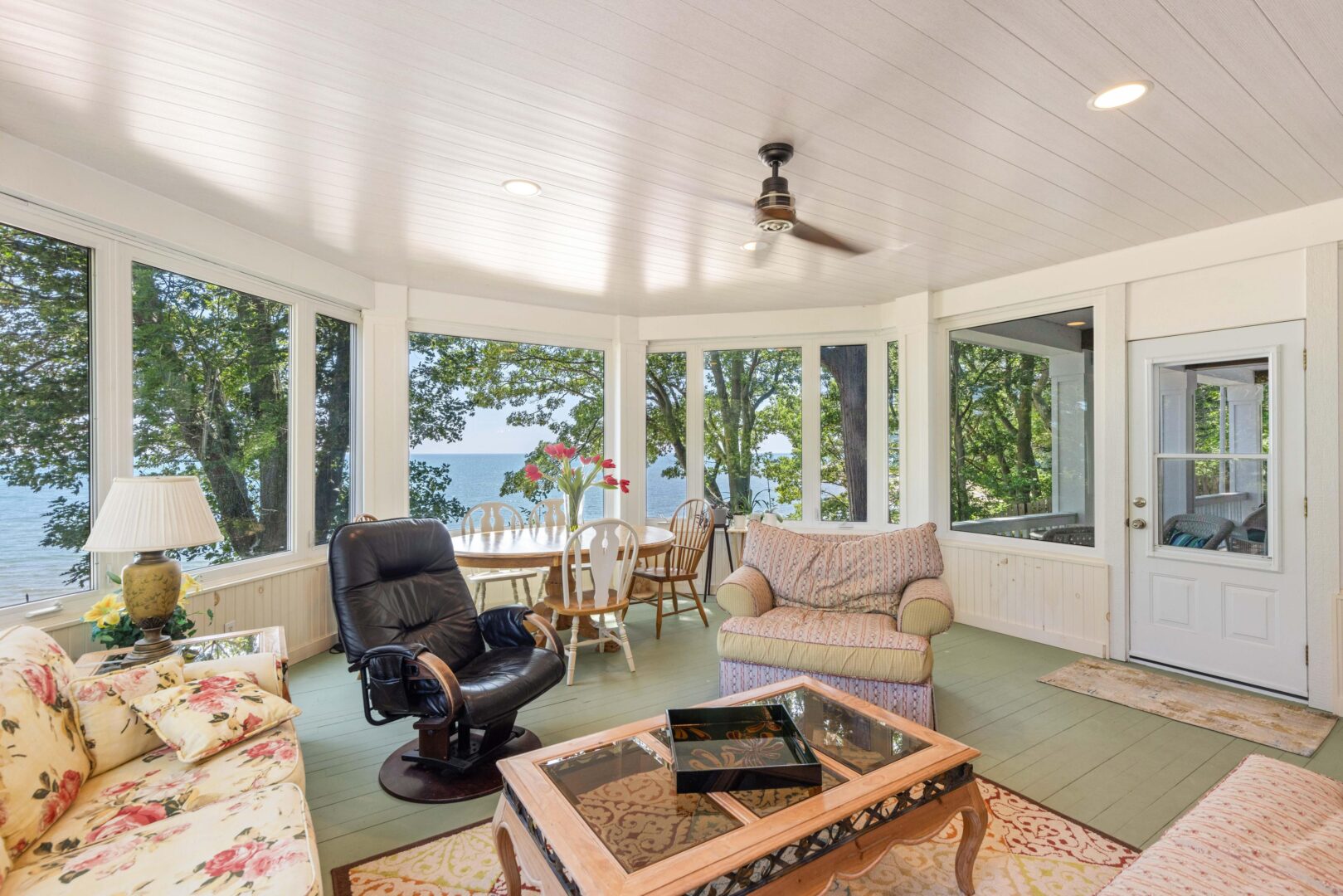 Bright sunroom with large windows, comfortable seating, and views of the lake through mature trees