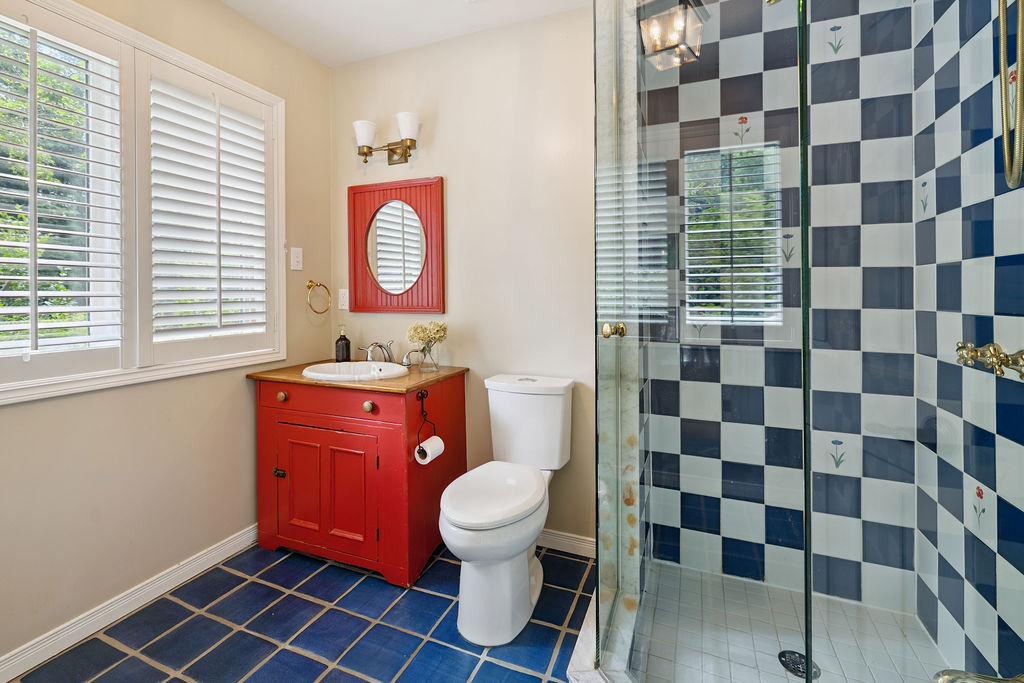 A white bathroom with blue tiled floors, a red vanity and checkerboard tiles in the glass shower