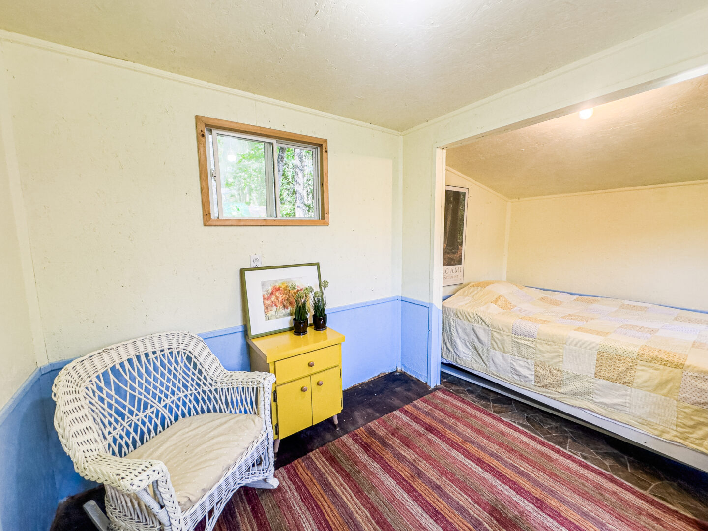 A small white room with a red striped carpet. To the left, a white whicker chair. A bed is tucked into a nook on the right