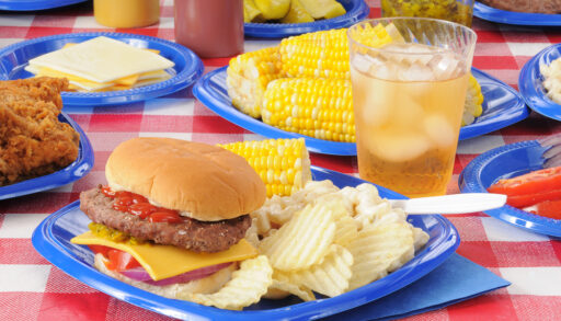 A cheeseburger with potato chips and corn on the cob on a picnic table loaded with food