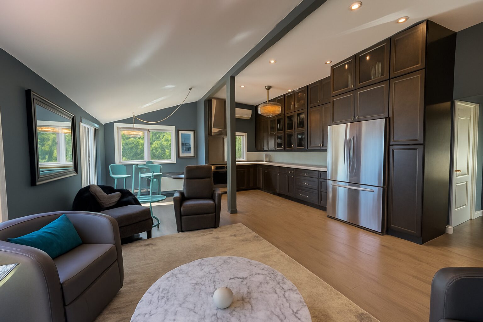 An open-concept living area with chairs around a low coffee table. To the right, a dark brown kitchen