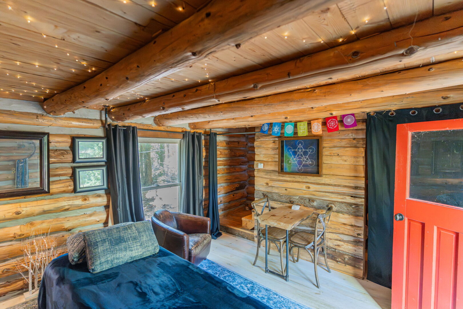 Log cabin interior with exposed beams, twinkle lights, red door, small dining set, and serene forest views
