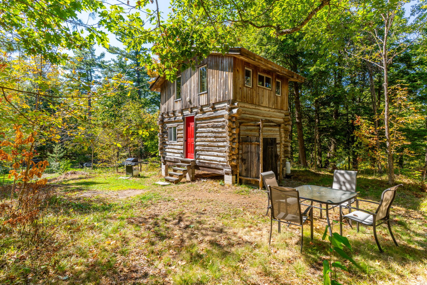 Rustic cabin with red door and upper-level windows, set in a clearing with BBQ and outdoor dining area among mature trees
