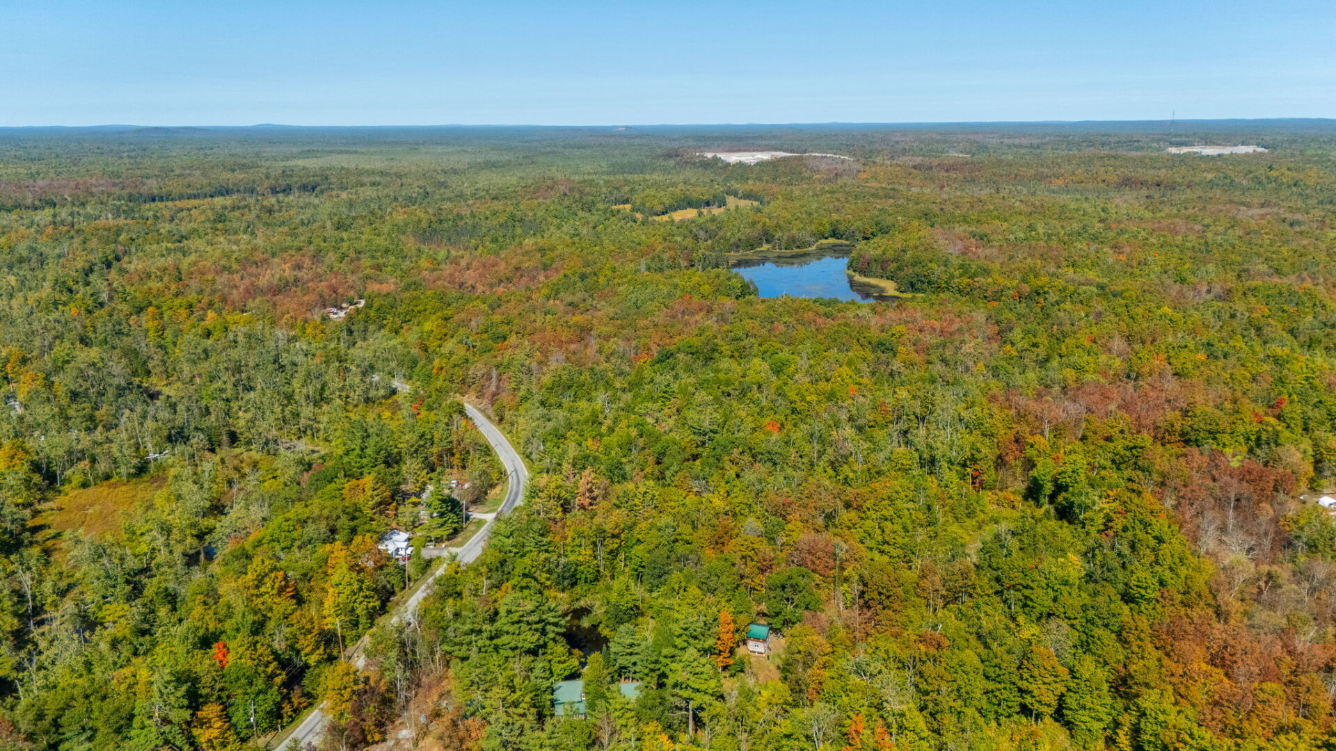 A wide drone shot showing a winding rural road through a dense autumn forest with a small lake in the distance, highlighting the seclusion and natural beauty of the property.