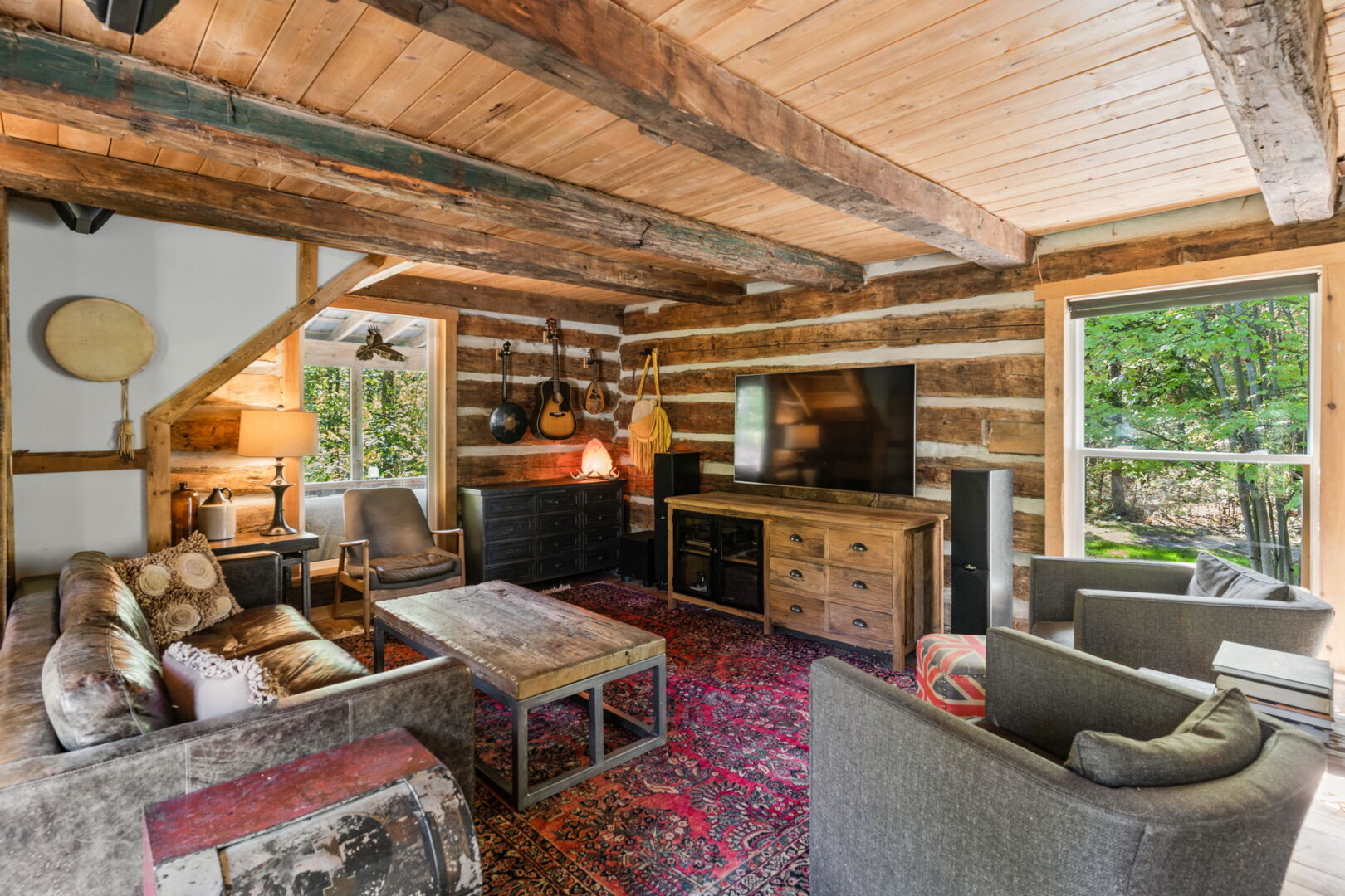 Warm and inviting living space with exposed beams, log walls, large windows, and eclectic decor including musical instruments