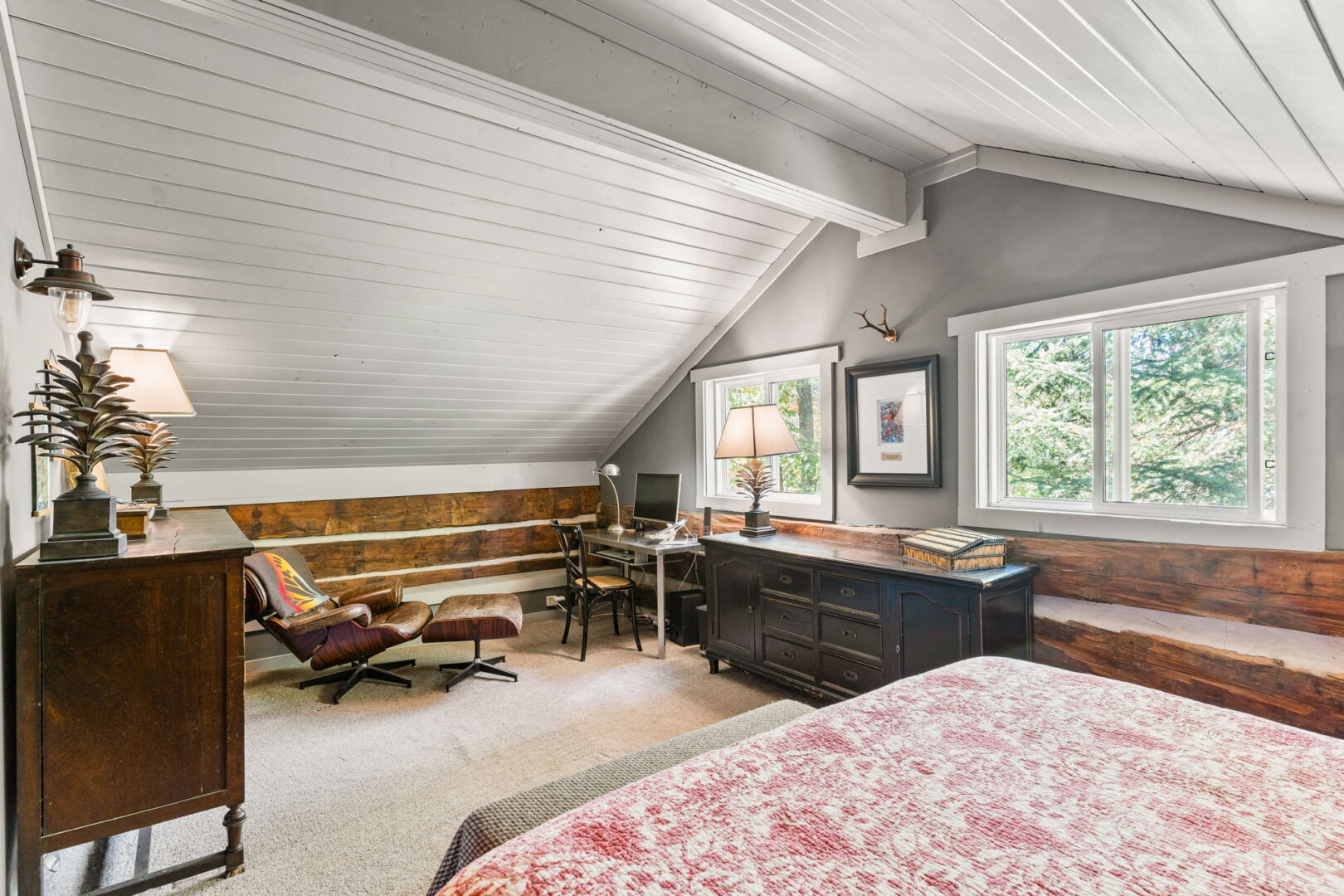 Upper-level bedroom with sloped ceiling, wood trim, large windows, and dedicated workspace overlooking the trees