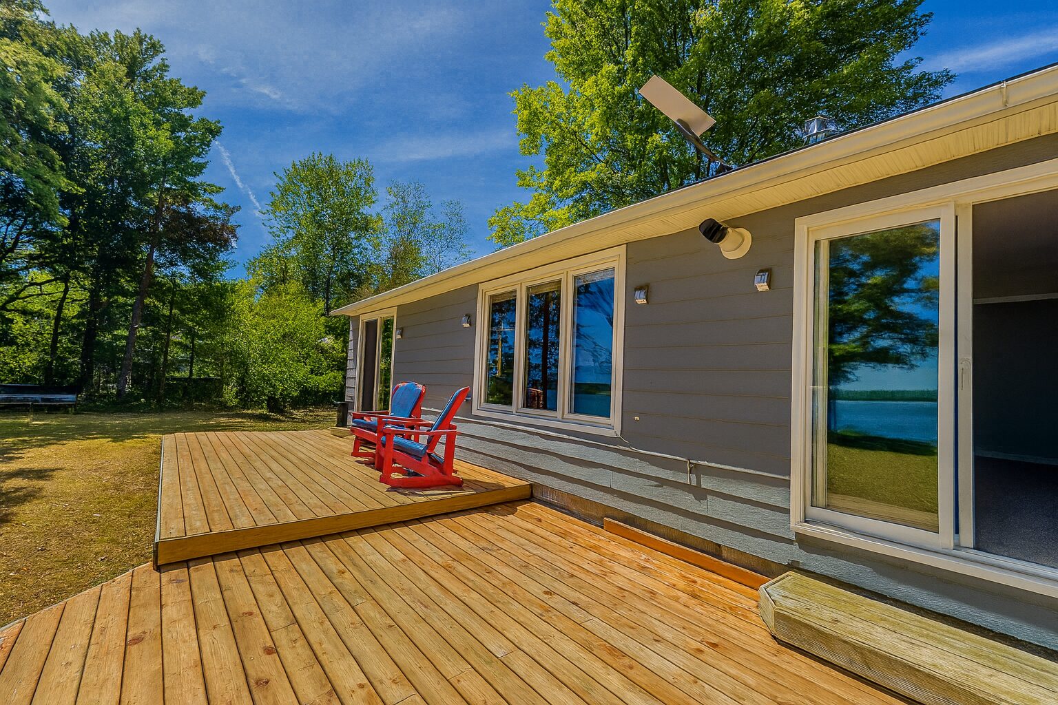 A wood deck with two red Muskoka chairs at the back of a grey bungalow cottage