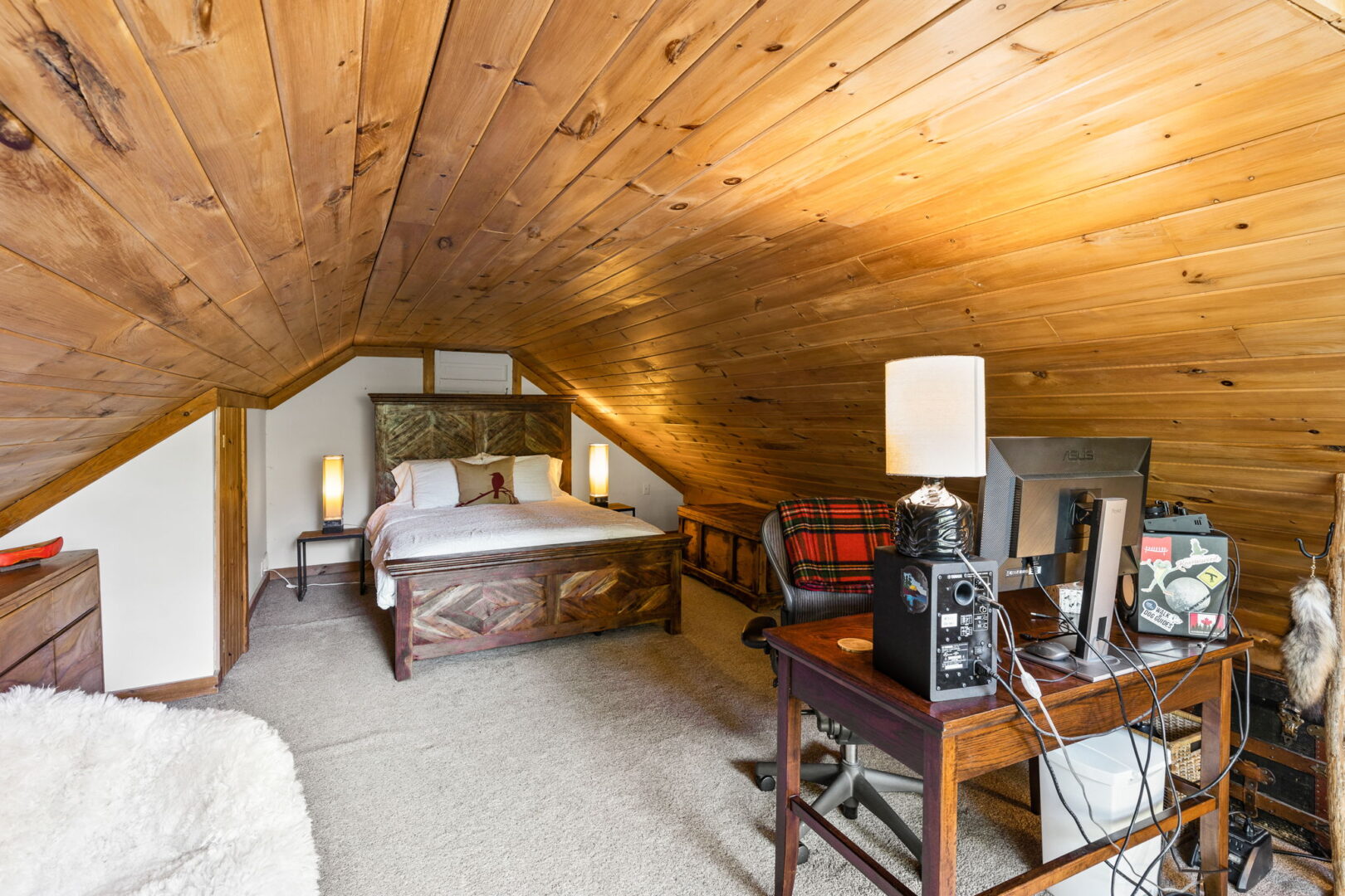 Attic bedroom with angled pine ceiling, wood bedframe, cozy carpet, and workstation setup