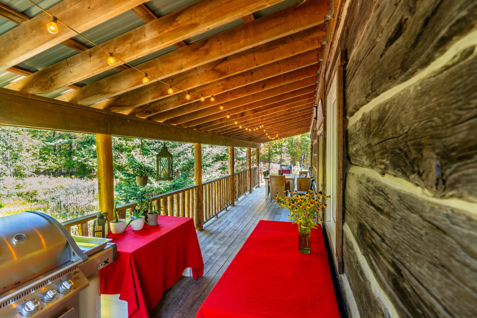 Rustic log cabin porch with wooden beams, red tablecloths, bistro lights, and a peaceful view of the surrounding woods