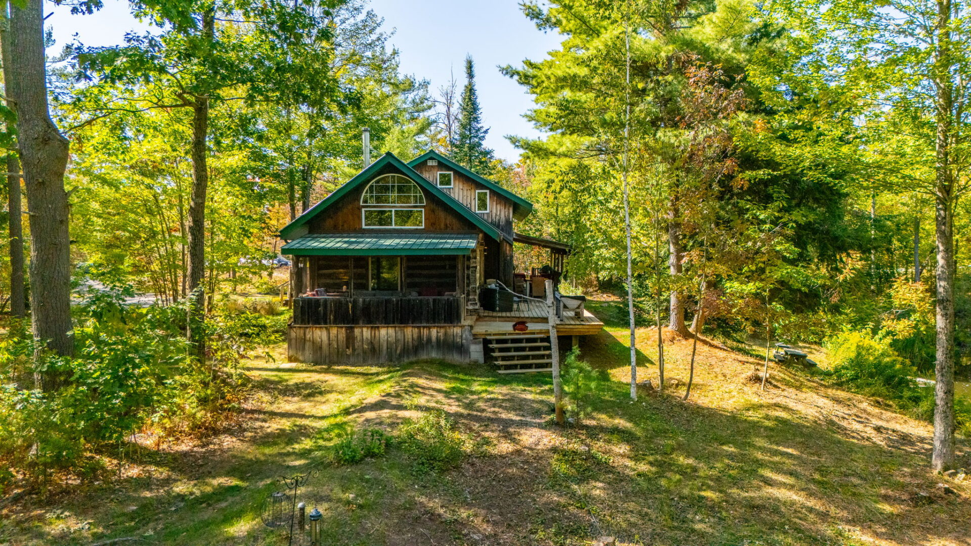 Back view of 2-storey log cabin with green metal roof, screened-in porch, and deck surrounded by trees and sloping lawn