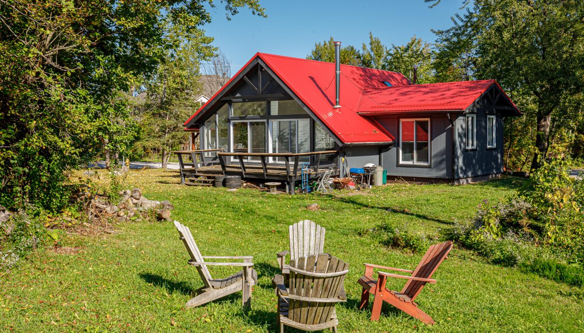 A large cottage with a red roof sits on a grassy lawn. In front, a fire pit area with four Muskoka chairs