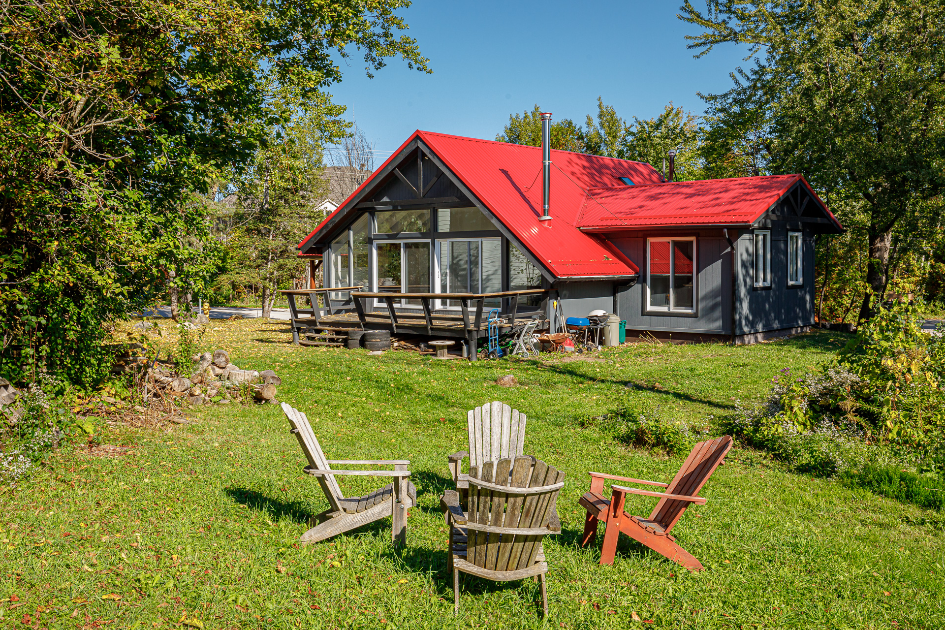 A large cottage with a red roof sits on a grassy lawn. In front, a fire pit area with four Muskoka chairs