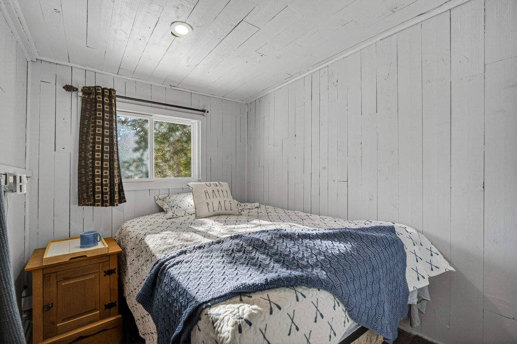 Simple bedroom with white-paneled walls, bed, side table, and natural light through the window.